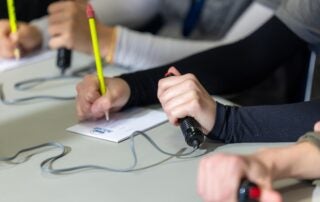Hands of students hold pencils over notepads and buzzer system handles.